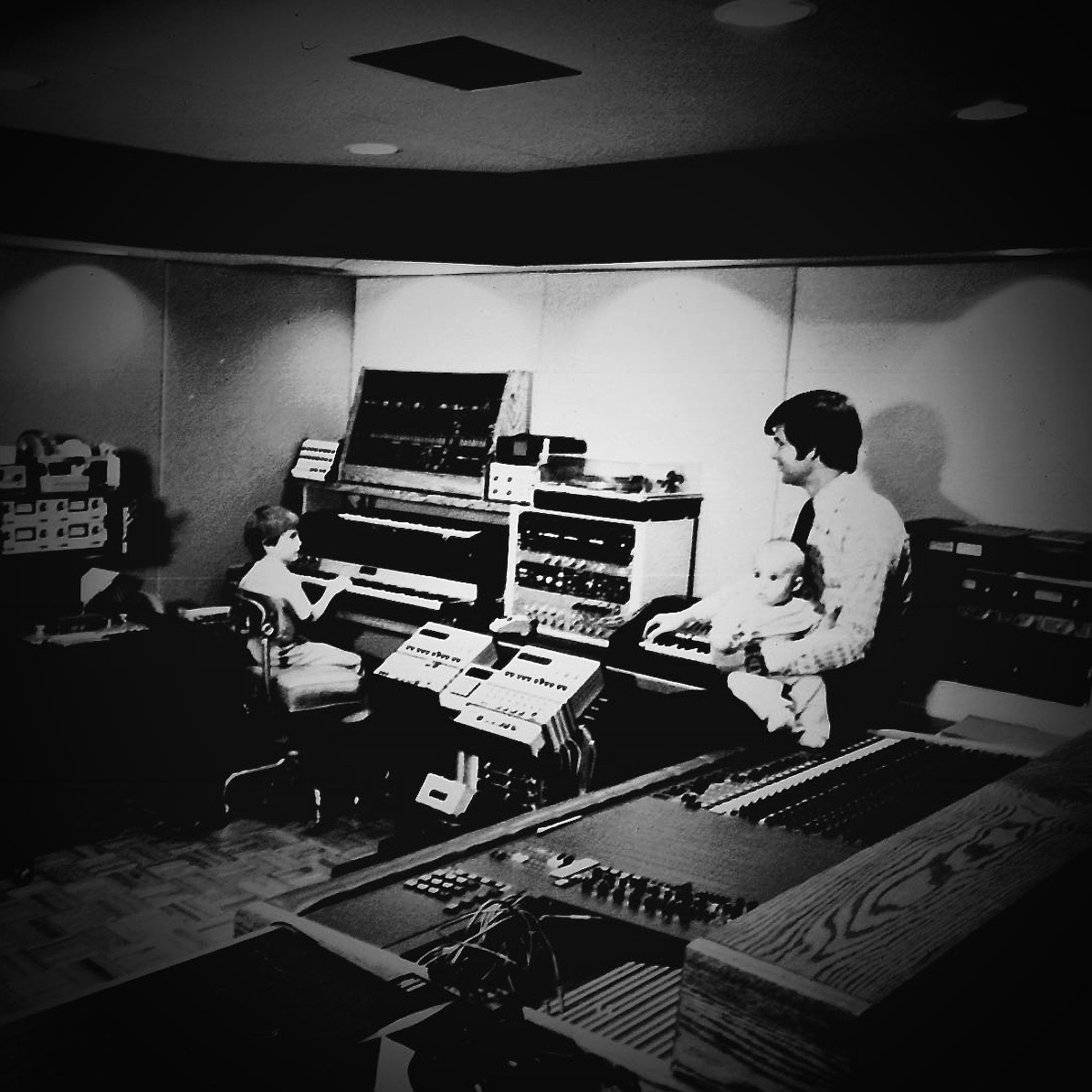 Archival black-and-white photograph of an early electronic music studio with an adult and two children at keyboards and synthesizer equipment